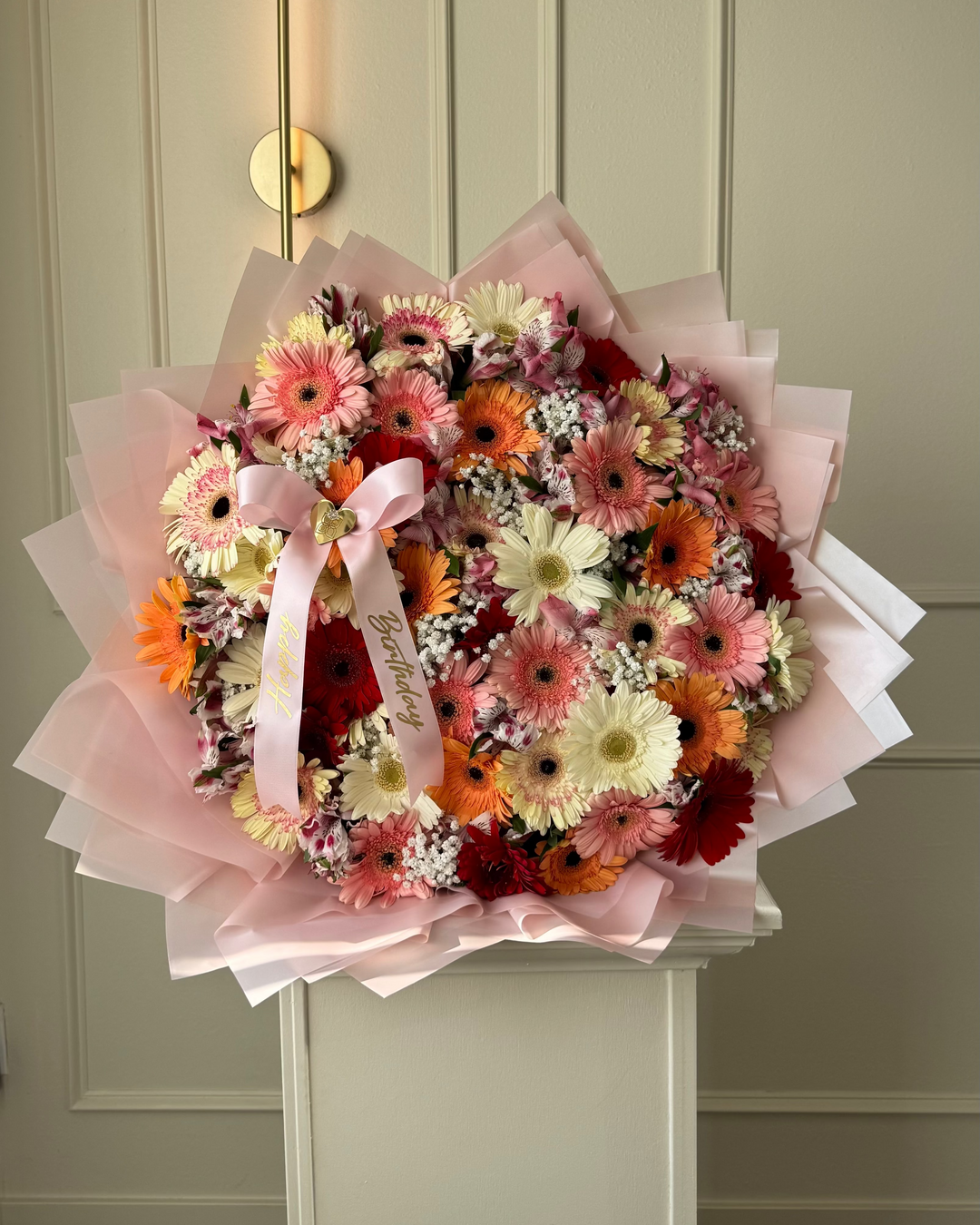 A large, vibrant circular flower bouquet featuring gerbera daisies in shades of orange, pink, white, and burgundy, wrapped in light pink tissue paper with a matching pink ribbon, displayed on a white pedestal in front of a beige paneled door.