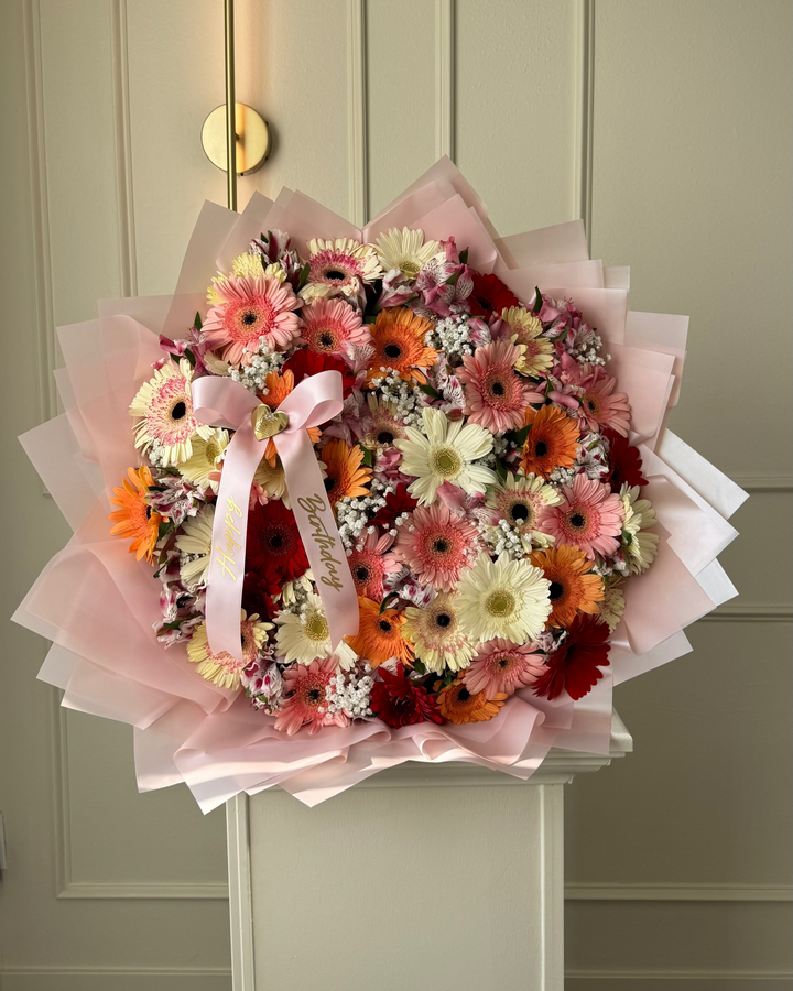 A large, vibrant circular flower bouquet featuring gerbera daisies in shades of orange, pink, white, and burgundy, wrapped in light pink tissue paper with a matching pink ribbon, displayed on a white pedestal in front of a beige paneled door.